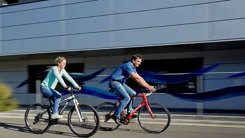 Zwei Menschen, die auf zwei Stadträdern vor einem Büro-Gebäude vorbeifahren Zwei Menschen, die auf zwei Stadträdern vor einem Büro-Gebäude vorbeifahren