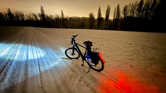 Ein beleuchtetes Fahrrad steht nachts auf einem verschneiten Feld; Scheinwerfer wirft blau-weißes Licht nach links, Rücklicht leuchtet rot nach rechts, im Hintergrund dunkle Baumreihe unter orangefarbenem Himmel.